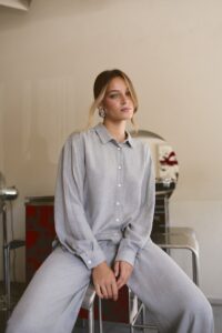 Szara koszula i szerokie spodnie rêver — siedzące ujęcie całej stylizacji na tle baru kawiarni